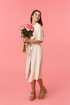 Full-length vertical portrait pretty brunette girl on romantic date receiving nice bouquet flowers, holding roses and gazing behind with delighted soft and tender smile, standing pink background.