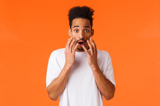 Scared, freaked-out and shocked african-american young hipster guy looking at something scary or frightening, standing concerned over orange background, hold hands near opened mouth.