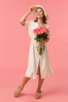 Full-length vertical shot gorgeous elegant woman in dress and hat, going home from date, holding beautiful flowers, received roses, smiling and feeling sunlight on her face, pink background.