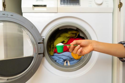 A woman's hand holds a cap with washing powder against the background of a washing machine with bright things put into it. The girl washes and wringes out things in the laundry room at home.