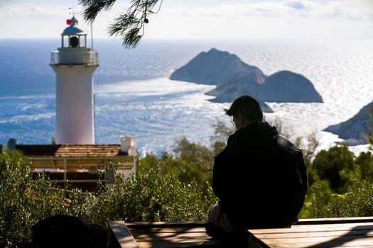 The traveler sits against the backdrop of a picturesque seascape with small islands and an old white lighthouse on the peninsula. A young man is trekking the paths along the sea.