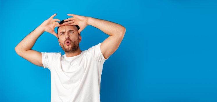 Portrait of a confused young man holding hands on his head over blue background, close up