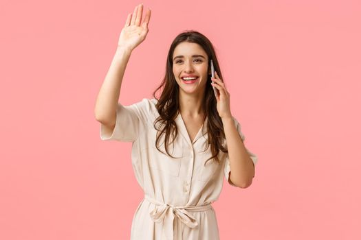 Cheerful attractive smiling caucasian woman seeing friend and waving hello, say hi as talking on phone, speaking, calling someone having conversation and greeting passerby, pink background.