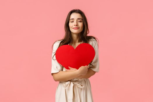 Care, tenderness and beauty concept. Tender and lovely young girlfriend in dress standing over pink background, hugging large heart card with closed eyes and cute smile, standing pink background.