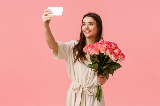 Birthday, holidays, romance concept. Charming alluring young caucasian woman receive delivery, got gift flowers, taking selfie on smartphone with beautiful bouquet, standing happy pink background.