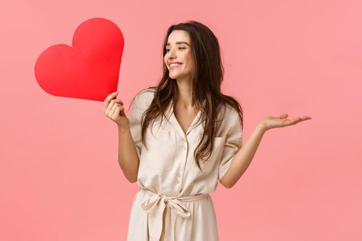 Surprises, relationship and romance concept. Attractive carefree and happy smiling brunette female in dress, raising hands amused and cheerful looking left, holding heart love card, pink background.
