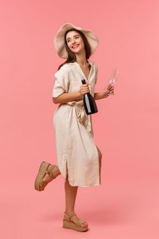 Full-length vertical shot woman on picnic, going out with girlfriend on fine sunny day in hat and dress, rushing with bottle champagne and glasses, smiling happily, standing pink background.