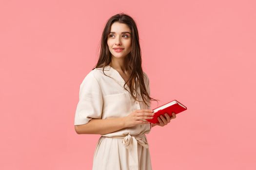 Attractive dreamy and romantic cute girl in dress turn around looking camera thoughtful, holding notebook, prepare notes for work, standing pink background, thinking. Copy space