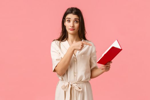 Did you wrote this. Intrigued brunette girl asking question about something written in notebook, pointing finger at note and looking puzzled, standing pink background confused. Copy space
