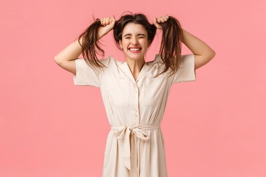 Emotions, celebration and leisure concept. Happy and funny cute girl in dress, messy hair and pulling it, jumping close eyes and smiling joyfully, having fun, fool around playfully, pink background.