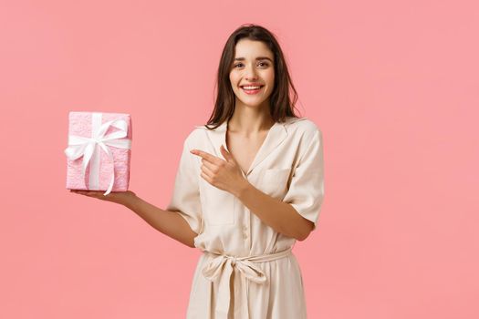Girl prepared romantic gift for anniversary. Cheerful pretty young woman in cute dress holding gift box and pointing finger at present with delighted, happy smile, standing pink background.