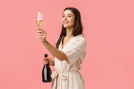 Girl raising glass for friend, give a toast looking left at person, holding champagne and smiling joyfully, standing over pink background, celebrating special occasion. Copy space