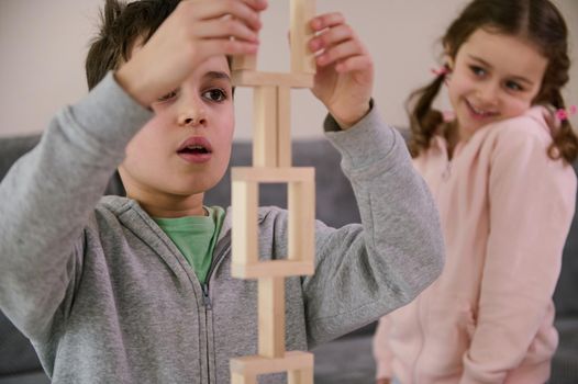 Close-up. Handsome schoolboy, concentrated on construction of complex tall structure from wooden blocks, against the background of his cheerful little sister, smiling sweetly, looking at his structure