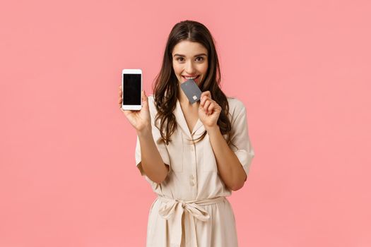 Romance, love and shopping concept. Cheerful carefree young woman order online, holding smartphone, biting credit card and smiling tempting, want buy new clothes, promote application, pink background.