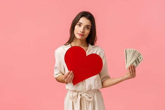 Decisions, relationship and life concept. Alluring romantic young woman having decision made, holding heart card near chest and showing money, tilt head smiling, making choice, pink background.