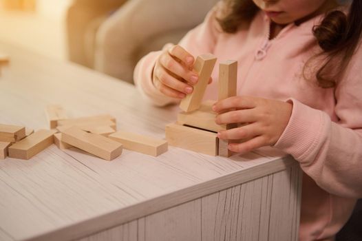 Close-up of little girl hands building wooden structure with blocks and bricks. Fine motor skills development, educational board games concept with copy ad space
