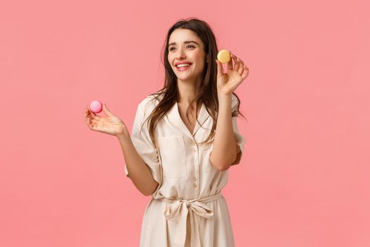 Pretty young female entrepreneur starting own business, baking desserts, suggest try friends, holding macarons and smiling joyfully, looking left amused, standing pink background delighted.
