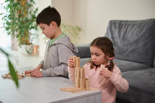 Creative Caucasian little girl sitting next to her brother and focused on construction of complex tall structure from wooden blocks, . Fine motor skills, concentration and educational leisure concept
