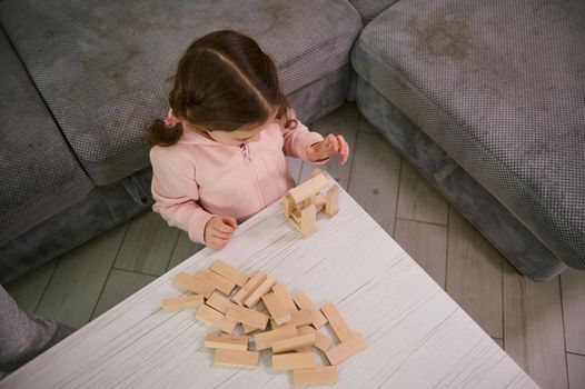 Overhead view of creative preschool child, concentrated baby girl on building with wooden blocks bricks. Educational board game and developmental smart pastime. Fine motor skill development concept
