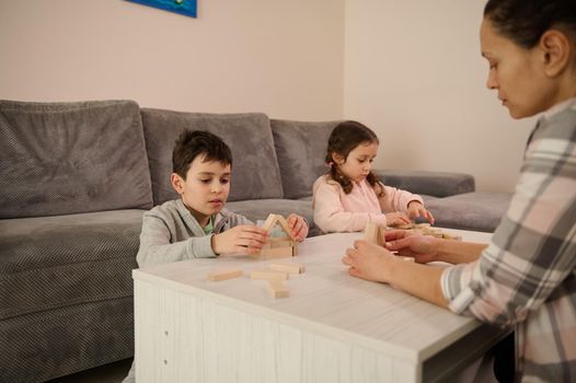 Soft focus on a handsome school age boy building wooden house with blocks and bricks, sitting at table near her sister and mom focused on construction of structures. Family pastime and leisure concept