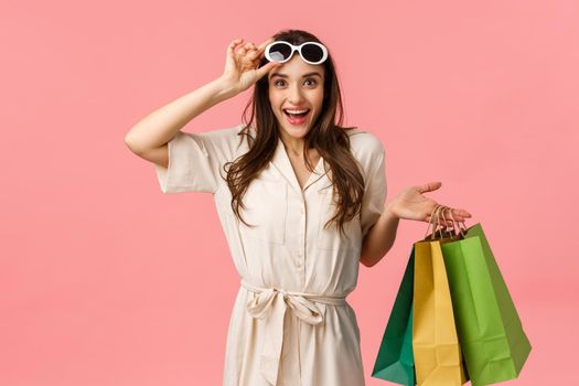 Excellent discounts, must have this season. Attractive young brunette in dress, holding shopping bags, taking-off glasses and smiling as seeing best price ever, standing pink background.