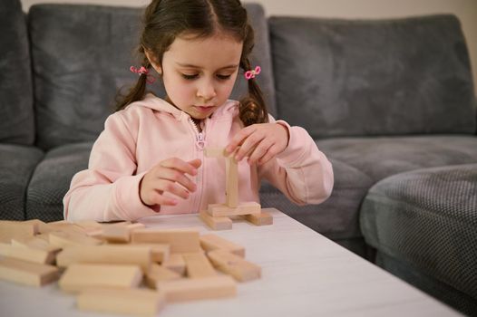 Adorable little girl with two pigtails, dressed in pink sweatshirt playing board game, building wooden constructions with blocks. Hand movement control and building computational skills concept.