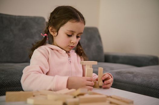 Cute adorable creative child European little girl playing board game, concentrated on building constructions with wooden blocks bricks. Hand movement control and building computational skills concept.