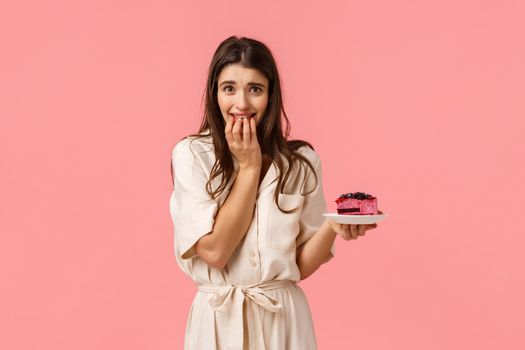 Temptation, diet and desserts concept. Alluring young feminine sporty brunette girl in dress, trying lose weight for summer, holding piece cake and biting fingers want eat but cant, pink background.