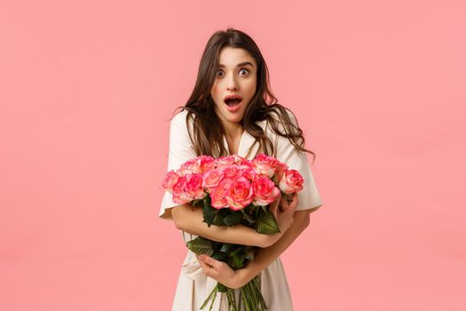 Surprised and excited attractive brunette girlfriend astonished by unexpected and amazing gift, secret admirer sent beautiful bouquet, gasping and looking impressed, pink background.
