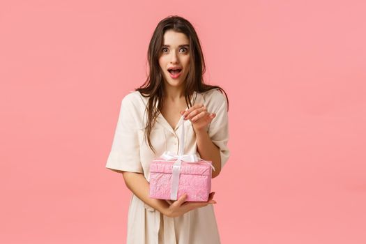 Excitement, tenderness and celebration gesture. Feminine surprised and excited alluring brunette girl in dress, wrapping gift, look amused and wondered, holding cute present box, pink background.