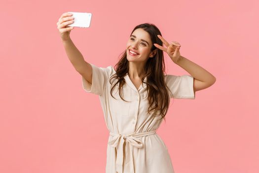 Carefree emotive, happy smiling brunette female in dress, holding smartphone, taking selfie and make peace sign, tilt head and smiling, sending positive vibes to followers, standing pink background.