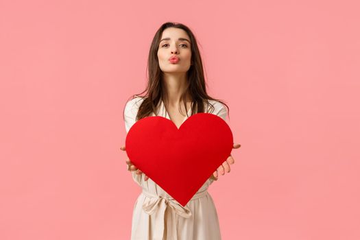 Tenderness, beauty and romance concept. Attractive tender and sensual young woman in dress, folding lips and blowing air kiss at camera as holding romantic heart card, valentines day.