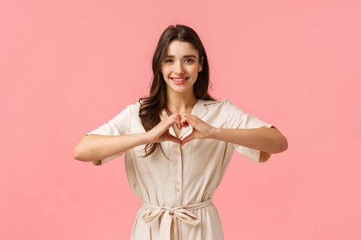 Waist-up portrait charming young brunette girl in dress, showing heart sign with love or care, absolutely adore boyfriend, having perfect relationship, feeling happy, smiling joyfully.