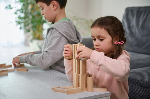Creative children, boy and girl playing with game stacking wooden toy blocks in high building structure. Hand movement control and concentration skills, educational leisure and board game concept