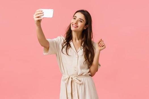 Beauty, fashion and women concept. Alluring young feminine woman taking selfie in new dress, posting new photo online, smiling tender and cute camera, standing pink background. Copy space