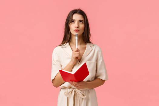 Thoughtful and creative young woman making list, pouting and looking up pensive and inspired, holding red notebook and pen, create new poem or prepare for exam, thinking over pink background.