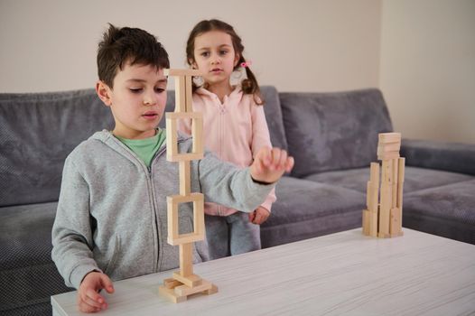 Focus on a handsome school aged boy building tall structure of many frames with wooden blocks while his little sister inspecting his construction. Children playing educational board game together