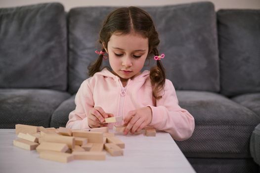 Education, development of fine motor skills, developmental board games concept. Adorable European girl concentrated on building with wooden blocks, sitting at a table in the living room
