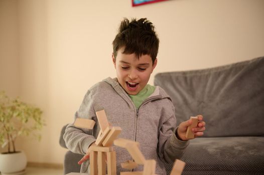 Close-up of a handsome school aged Caucasian boy playing board games and looks surprised when his constructed structure from wooden blocks falling down. Education leisure and board games concept