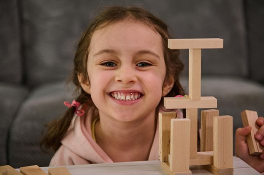 Close-up portrait of a beautiful adorable little girl smiling toothy smile, looking at camera, enjoying educational board game, building wooden structures with blocks. Development of fine motor skills