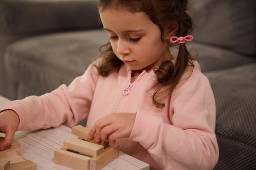 Close-up portrait of charming adorable little preschool Caucasian girl in pink sweatshirt playing with wooden blocks, building structures. Fine motor skills development and educational leisure concept