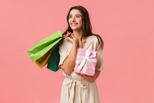 Lifestyle, holidays and emotions concept. Cheerful carefree young feminine woman holding shopping bags and wrapped cute gift, looking aside smiling, standing pink background delighted.