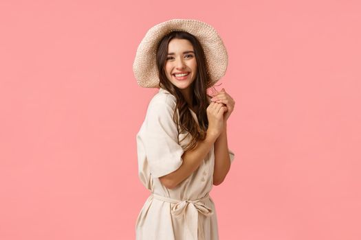 Feminine, coquettish and flirty woman with dark curly hair, wearing trendy hat and stylish dress, laughing, enjoying beautiful romantic date, clap hands and gazing camera delighted, pink background.