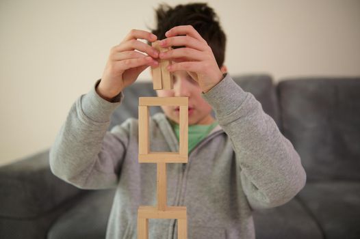 Close-up of Caucasian handsome puzzled school boy's hands, concentrated on construction of complex tall structure from wooden blocks. Fine motor skills, concentration and educational leisure concept