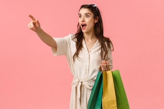 Woman seeing wonderful dress in store on manequin. Attractive cheerful young female in dress, holding shopping bags pointing finger sideways and gasping amazed, standing pink background.