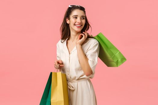 Relaxed and carefree glamour, feminine cute girl in dress holding shopping bags and turning right with beautiful happy smile, enjoying shop trendy stores, standing pink background. Copy space