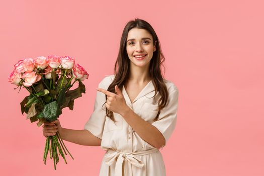 Presents, valentines day, beauty concept. Gorgeous feminine girl florist made beautiful bouquet for customer, holding roses and pointing finger at flowers, smiling silly and happy, pink background.
