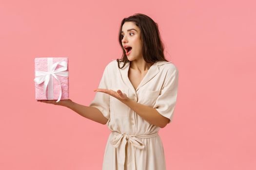 Girl likes surprises. Amused and happy cheerful young brunette woman in dress, receive gift box, pointing at present looking impressed and astonished as didnt expect such cuteness, pink background.