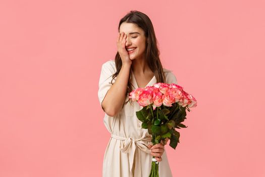 Happiness, love and relationship concept, Woman feeling cherished and valued, attractive brunette girl in stylish dress, holding roses, flower bouquet and smiling, laughing shy, pink background.