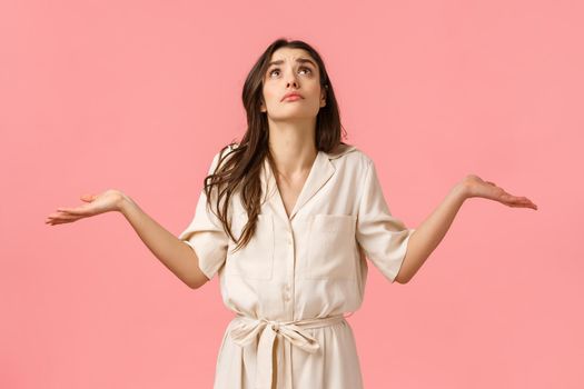 Girl cant understand what faith prepared. Confused and indecisive glamour young brunette girl in dress, shrugging with hands sideways, looking up pouting rain, pink background. Copy space